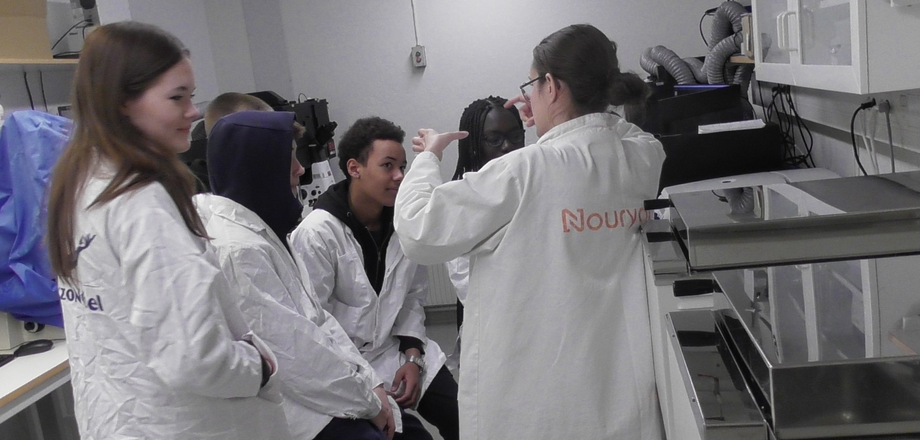 Fem glada ungdomar i vita rockar som lyssnar på en engagerad vuxen forskare.