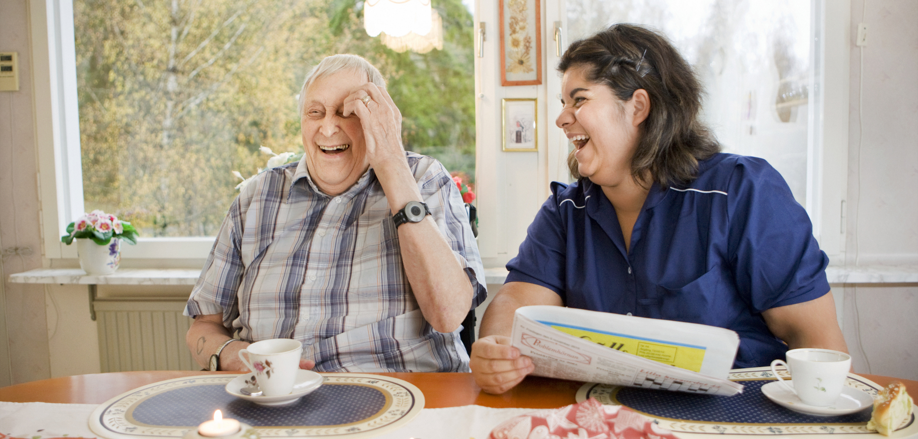 Äldre man och hemtjänstpersonal skrattar vid fikabordet.